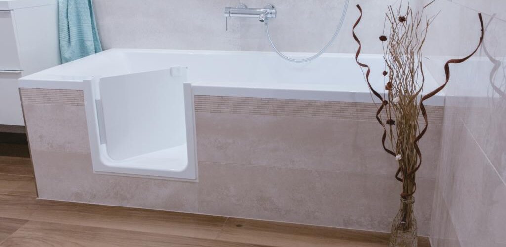 Eine moderne Badewanne mit geöffneter, barrierearmer Tür in einem stilvoll eingerichteten Badezimmer. Eine Vase mit dekorativen Zweigen und eine minimalistische Einrichtung ergänzen das Design.